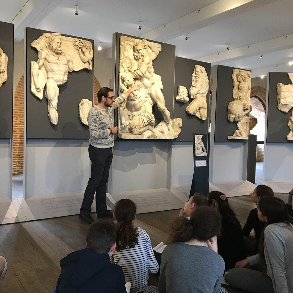 Un groupe d'élèves, assis devant les reliefs des travaux d'Hercule, assiste à une visite guidée conduite par un médiateur culturel.