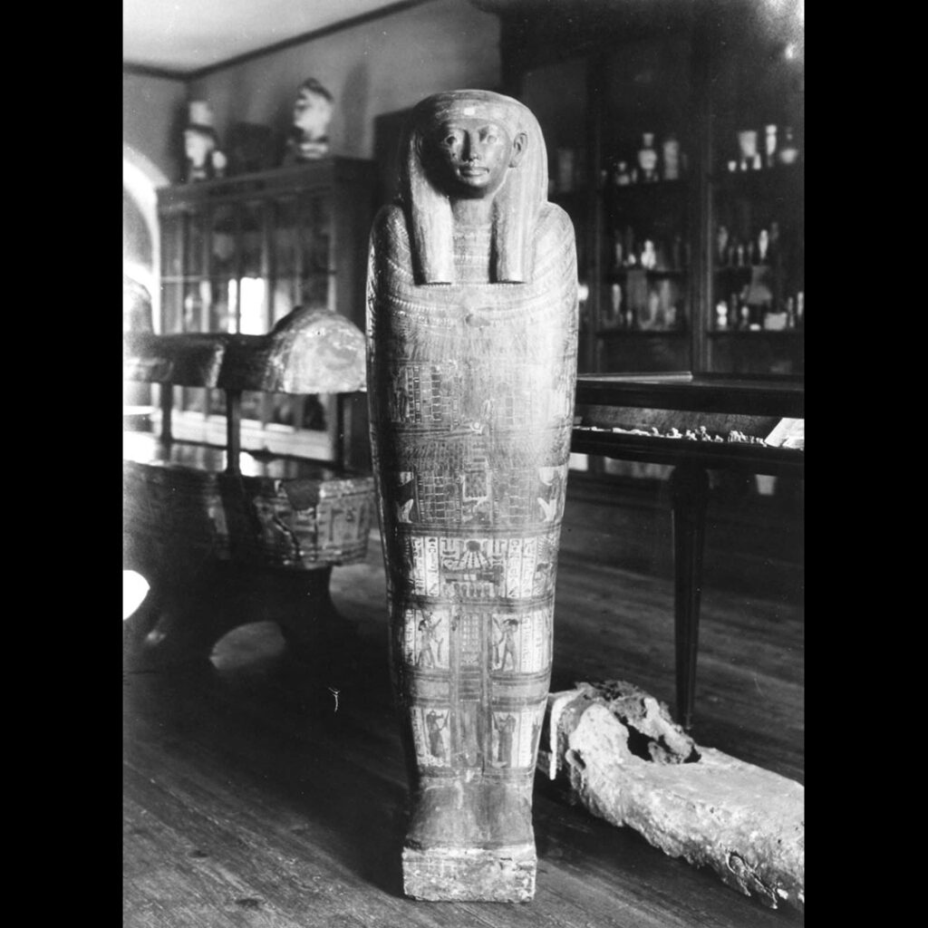 Photographie d'une ancienne présentation de salle au musée Saint-Raymond. Devant, on voit un sarcophage égyptien présenté debout. Derrière lui, d'autres sarcophages sont présentés allongés en compagnie de vitrines contenant de petits objets. Derrière, d'autres vitrines exposent des objets divers, des poteries ou des sculptures.