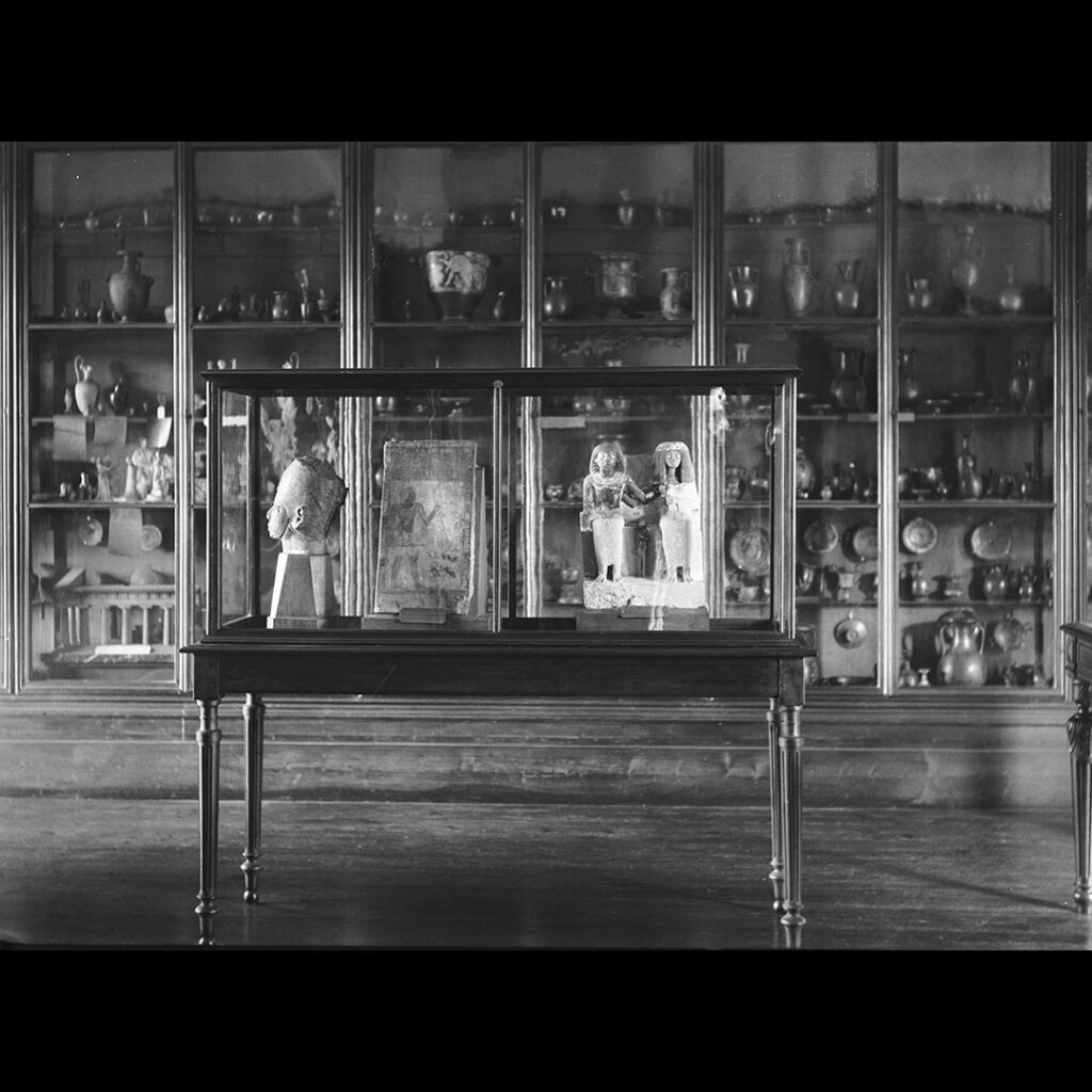 Photographie d'une salle du musée Saint-Raymond. Devant, une vitrine au milieu de la pièce expose des statues égyptiennes. À gauche, une tête en pierre. Au centre une peinture sur bois et à droite, un couple égyptien assis. Derrière la vitrine, contre le mur, d'autres vitrine allant jusqu'au plafond conservent des céramiques grecques et une maquette d'un temple grec.